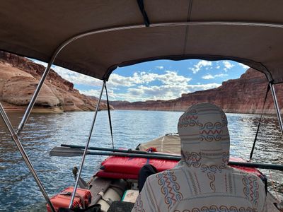 Seen from under the boat's tan bimini, a crew member in a patterned white hooded sun shirt sits facing forward past stowed oars, red gear rolls and an orange hard case, with glassy reservoir water and red canyon walls marked by a pale waterline ahead.