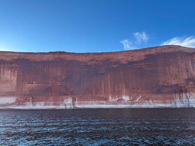 A long sheer red sandstone wall streaked top to bottom with dark desert varnish rises above a broad bleached white waterline band, with dark wind-rippled reservoir water across the foreground under a deep blue sky.