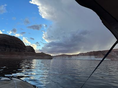 From under the boat's canopy, heavy gray clouds trailing rain hang over glassy reservoir water at dusk, with a rounded slickrock dome on the left and a long bleached waterline band along the far shore catching the last light.