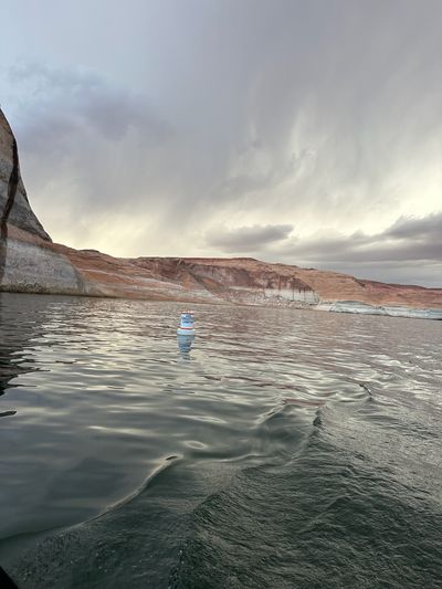 A white and orange navigation buoy lettered FORGOTTEN CANYON floats on gray reservoir water with the boat's wake curling through the foreground, below red cliffs banded by a bleached waterline under a rain-streaked dusk sky.