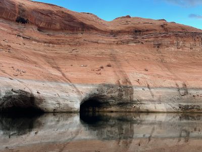 A dark cave-like alcove is eroded into the bleached gray band at the base of a red slickrock slope and mirrored in still reservoir water, with loose cobbles scattered up the sandstone above under a blue sky.