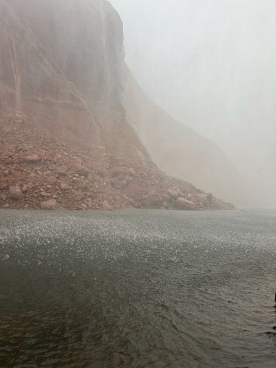 Heavy rain pocks the gray-green water across the foreground while a red talus slope and the cliff wall behind it fade into storm haze.