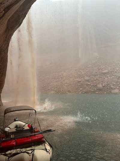 A muddy, sediment-loaded waterfall pours off the rim past the lip of a sandstone overhang beside a rigged raft with a tan bimini, red gear rolls and an orange case, while heavy rain hammers the green water and a rubble slope fades into mist beyond.