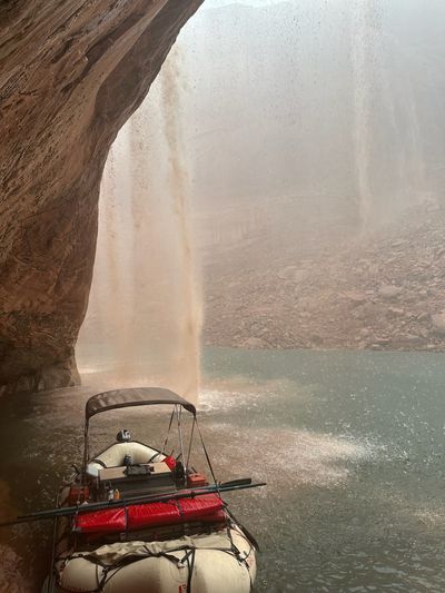 The same muddy waterfall drops off the overhang lip beside the moored raft, with a second fainter fall streaking the misted wall to the right and rain dimpling the green water across the whole frame.