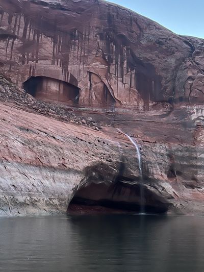 A thin fresh waterfall drops over the bleached band of a red slickrock slope into still water, below dark varnish drip streaks and two large shadowed alcoves cut into the wall above; the frame is slightly soft.