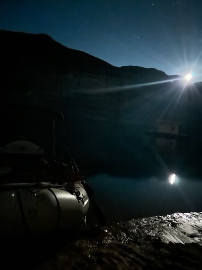 A rising moon burns a starburst over a dark ridge and lays a silver path across glassy black water, with the pale tube of a moored raft on a wet rock shelf in the foreground and stars scattered overhead.