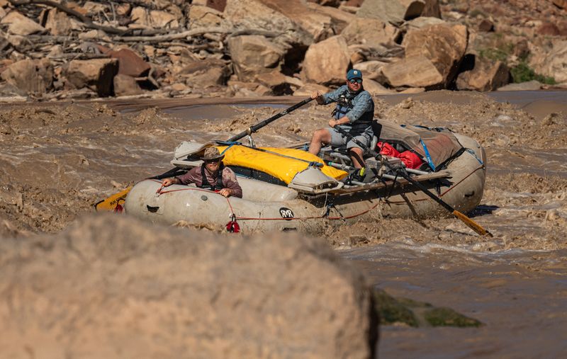 A loaded white gear raft runs brown whitewater: an oarsman in a teal cap and black life jacket pulls on the oars