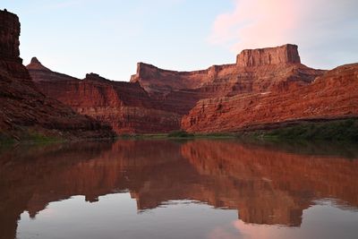 Glassy brown river mirroring tiers of red ledged cliffs, a pointed peak and a flat-topped mesa in soft evening light, pink cloud in a pale sky and a green riparian strip at the waterline.