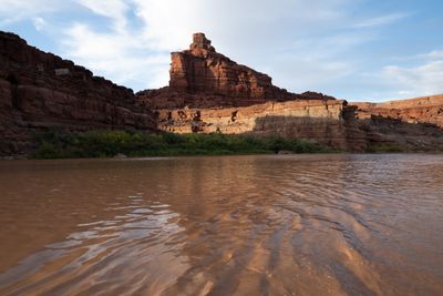 Low view across rippled, silt-brown river toward a stacked red sandstone tower and ledged cliffs, late sun lighting the lower wall above a green riparian bank, blue sky with wispy cloud.