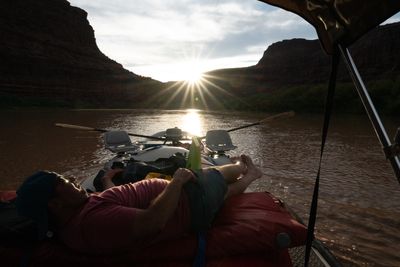 From under a raft's canopy, a man in a cap and sunglasses reclines on red bags holding a drink; ahead a gray raft with swivel seats and oars, and the sun bursts through a canyon notch over brown water.