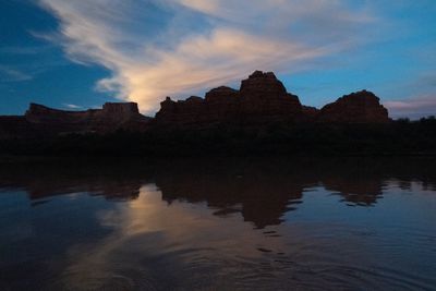 Stepped sandstone buttes and a flat-topped mesa silhouetted at blue hour, a plume of pink-lit cirrus above them, all reflected in dark, gently rippled river water.