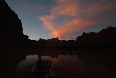 An oarsman silhouetted at the oars of a raft heads down a dark, still river between black canyon walls toward a flat-topped mesa, orange-lit clouds streaming up into a blue dusk sky.