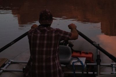 Seen from behind, a man in a plaid shirt and floral cap pulls on a pair of oars from a raft's frame seat, an orange dry box beside him, red cliffs reflected in the dim river ahead.