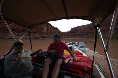 Two men laugh under a raft's tan canopy, one in a sun hoodie and trucker cap, one in a pink shirt holding a can on red bags; a gray raft with oars rides ahead on a brown river between red cliffs.