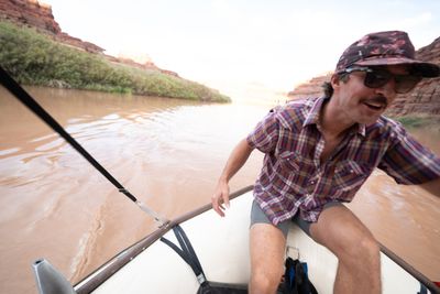 A man in a plaid snap shirt, floral cap and sunglasses crouches on the white tube of a raft, grinning, as the boat motors up a wake on a brown river below willow-lined banks and red cliffs.