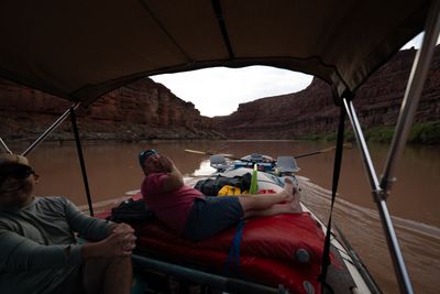Under a tan canopy on a raft, one man laughs at left while another in a pink shirt lounges barefoot on red bags and hollers ahead; a gray raft leads up a brown river between tall red walls.