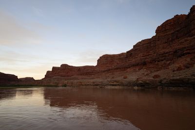 Calm, brown river curving past long red ledged cliffs in fading evening light, the walls reflected in the smooth water under a pale sky.