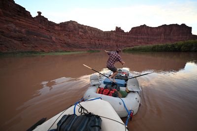A barefoot man in a plaid shirt balances on a cooler across a loaded gray oar-rigged raft lashed ahead of the camera raft, on smooth brown water below tall red walls with a balanced rock on the rim.