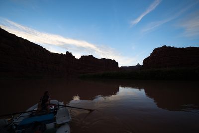 Silhouetted canyon walls with a spire and a distant pointed peak against a blue evening sky; in the lower corner a man at the oars of a gray raft dips a blade into still, reflective water.