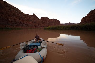 A smiling man in a plaid shirt and cap rows a loaded gray raft lashed ahead of the camera boat, oars spread over smooth brown water below tall red sandstone walls and a distant pointed peak.