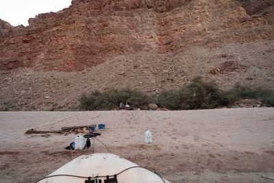 From the bow of a raft tied up on a wide sand beach, water jugs and gear sit on the sand as a few people gather at a camp under green trees at the foot of a talus slope and red cliff.