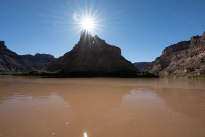 The sun bursts into a starburst over the top of a dark, backlit butte, with calm muddy brown water reflecting it in the foreground and canyon walls on either side.