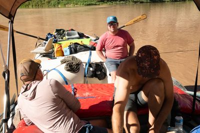 Under a bimini on a lashed-together raft rig, a man in a pink tee and blue cap leans on a loaded gear raft and smiles, as two others sit and crouch in the foreground on muddy water.