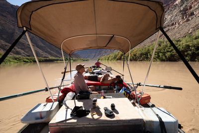 From the back of a motor raft under a tan bimini, a man in a cap sits and another lies shirtless on a red pad, while a rower in a red shirt works the oars of the lashed raft ahead down a muddy river between canyon walls.