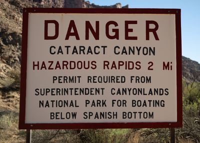 A white sign with a red border reads "DANGER, CATARACT CANYON, HAZARDOUS RAPIDS 2 Mi, PERMIT REQUIRED FROM SUPERINTENDENT CANYONLANDS NATIONAL PARK FOR BOATING BELOW SPANISH BOTTOM" in front of brush and canyon slopes.