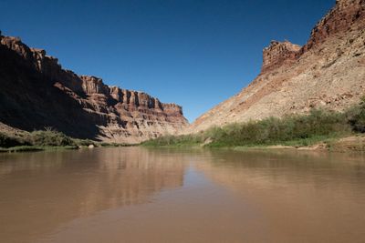 Calm muddy brown water reflects layered red and tan canyon walls, one side in shadow, that narrow toward a V-shaped gap under a deep blue sky.