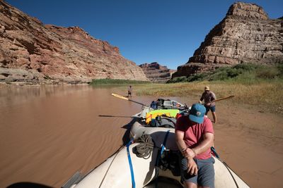 View from a gear raft's bow: one man wades knee-deep near a grassy mud flat while another holds an oar on the flat, and a man in a blue cap sits on the tube, between red and tan canyon walls.