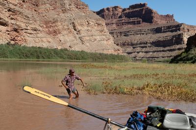 A man in a plaid shirt and cap wades knee-deep in muddy brown water beside a flat of grass and tangled vegetation, an oar blade and loaded raft deck in the foreground, red cliffs above.