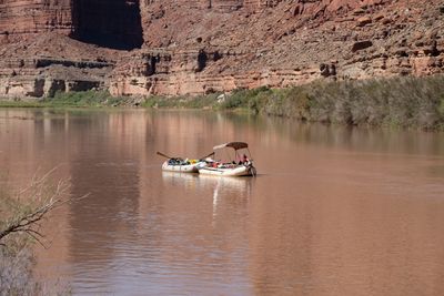 Two loaded gear rafts lashed side by side float on flat, muddy brown water as one person in a red shirt works the outboard under a tan bimini, below red ledgy cliffs and riparian brush.