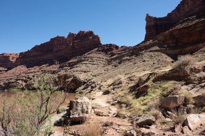 A dusty trail climbs a sunlit desert slope of boulders, grass and shrubs beside a muddy river, with two small hikers ahead and red sandstone cliffs against a clear blue sky.