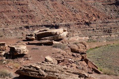 Weathered tan sandstone slabs sit on a red dirt rim above a sage-covered bench and a sliver of muddy river, with layered red slopes behind.