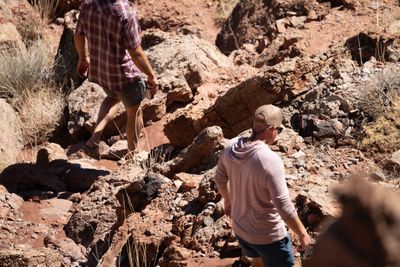 Two hikers seen from behind pick their way down a rough path through broken red rock and dry grass, one in a plaid shirt and one in a cap and pink hooded shirt.