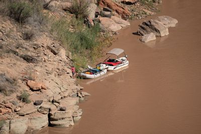 From a slope above, two loaded gear rafts, one under a tan bimini, sit tied to a sandstone and sand shore on muddy brown water as one person in a red shirt stands beside them.