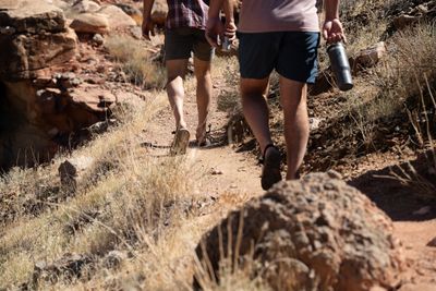 Close, low view of two hikers' legs in shorts and sandals walking a red dirt trail through dry grass and rocks, one carrying a steel water bottle and the other a cup.