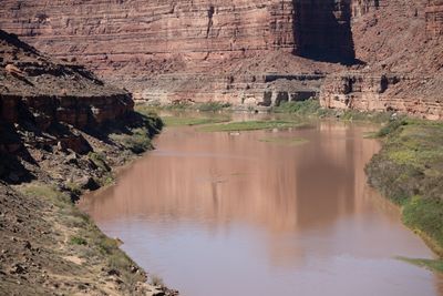 From a slope high above, a muddy brown river curves between grassy banks and small vegetated bars, a tiny raft on the water, under tall red layered sandstone walls.