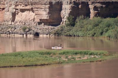 A loaded double rig of two rafts, one under a tan bimini, floats on muddy brown water beyond a grassy island, below pale and pink sandstone ledges and green riverside trees.