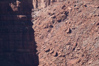 Telephoto view of a shaded red sandstone cliff edge beside a sunlit slope of broken red rock and boulders.