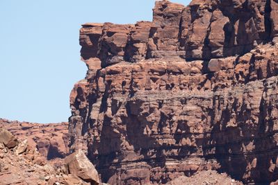 A sunlit red sandstone cliff of stacked, blocky ledges and darker thin beds rises against a pale blue sky, with broken boulders at its base.