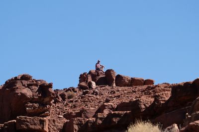 A lone hiker in a cap and plaid shirt sits atop a pile of red sandstone boulders on a ridgeline under a wide, cloudless blue sky.