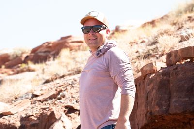 A smiling man in a tan trucker cap, dark sunglasses and a pink hooded sun shirt looks back over his shoulder on a sunny, rocky desert slope.
