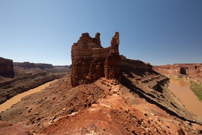A red sandstone tower with twin pinnacles rises from a rocky ridge between two stretches of muddy river in deep canyons, under a broad clear blue sky.