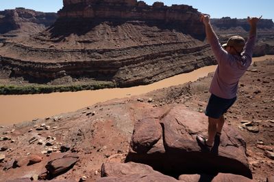 A hiker in a cap and long-sleeve shirt stands on a red boulder with both arms raised, throwing shakas over a muddy river curving below layered canyon slopes.