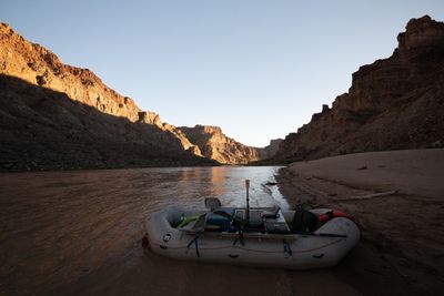 A gear raft with an oar frame and stowed oars sits beached on a sand shore in calm muddy water, as late sun lights the upper canyon walls downstream under a pale evening sky.