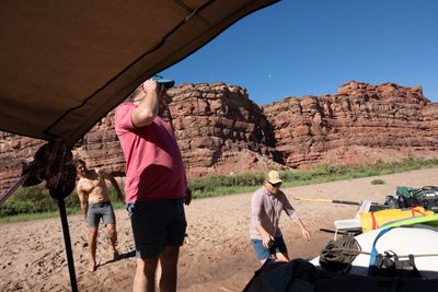 Three men in shorts on a wide sand beach load a beached gear raft under a tan bimini, red ledgy sandstone cliffs and a daytime moon in a clear blue sky behind.