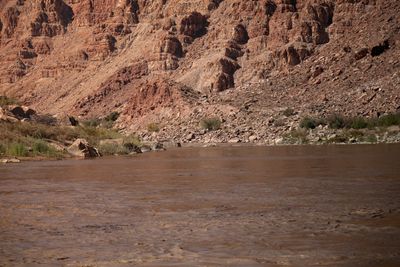 Low view across smooth, boiling brown river water toward a rocky shore and talus slope below red-brown ledged cliffs.