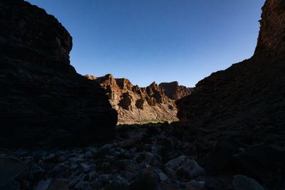 From a shaded boulder-strewn wash, dark canyon walls frame a gap onto sunlit ledged cliffs across the river corridor under a clear sky.